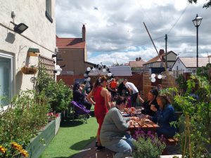 People sitting in a garden, some eating food, others wandering around.